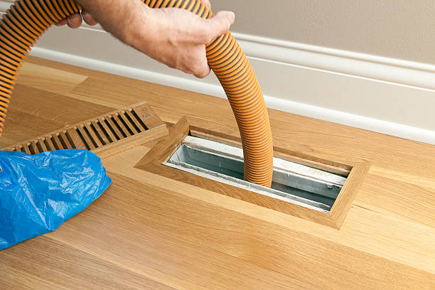 A worker is using a shop vacuum to clean a vent and duct on a new hardwood floor. The blue plastic is a protective cover over the worker's shoe.